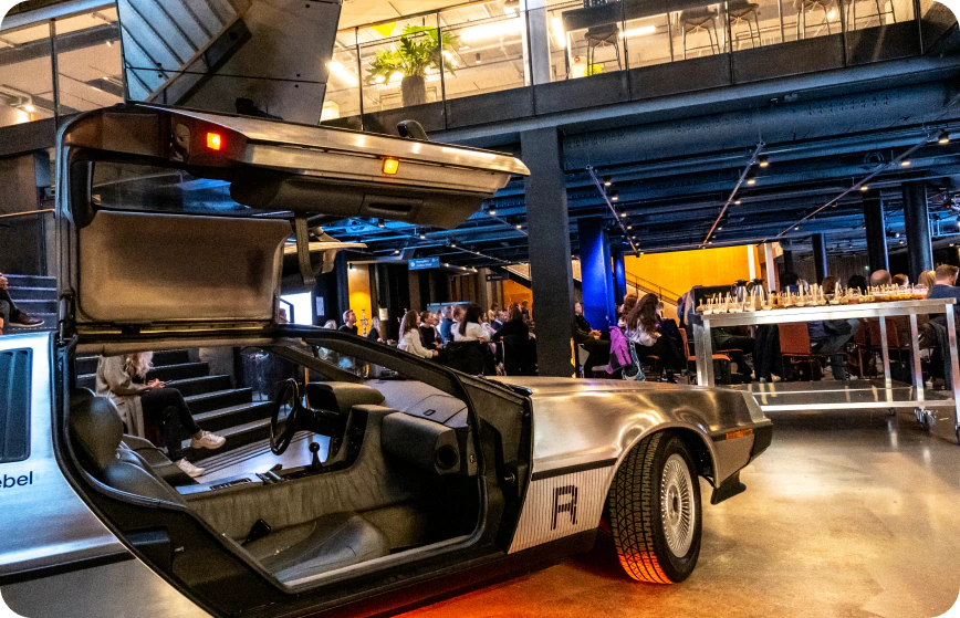 Event in progress at a Rebel space. A silver car is on display in the foreground. A large audience sits in the background.